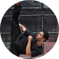 Man performing a high kick in black outfit outdoors on a sunny day.