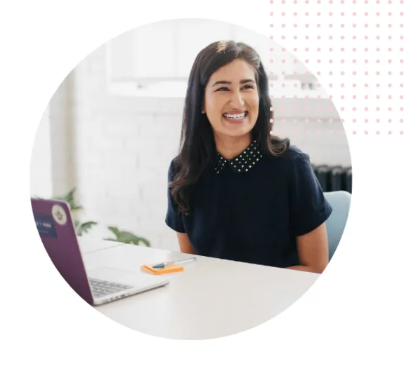 Woman smiling at a desk with a laptop and notepad in an office setting.